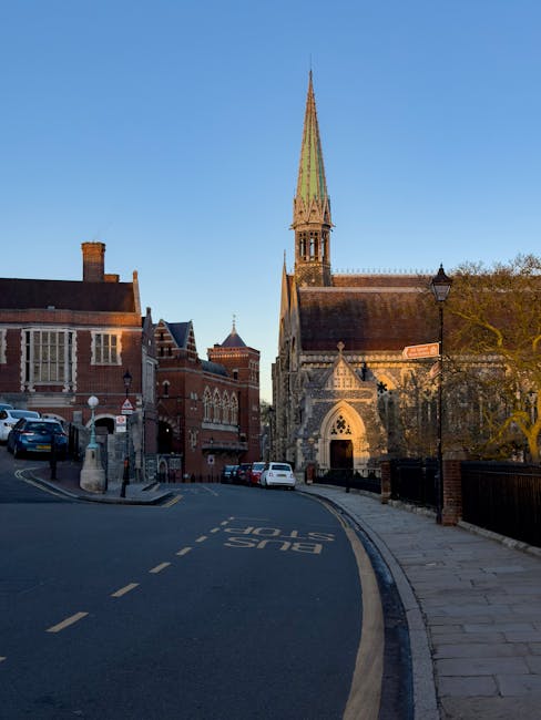 A view of a narrow street in Harrow with a church featuring a tall, pointed spire in the background. The street is lined with brick and stone buildings, some with decorative windows and brick chimneys