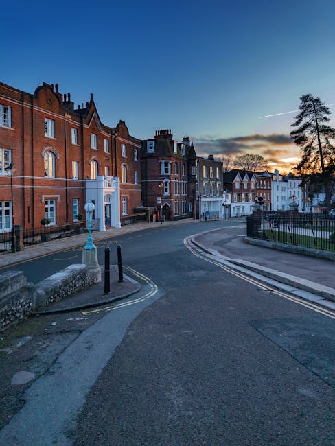 A view of a narrow street in Harrow with a church featuring a tall, pointed spire in the background. The street is lined with brick and stone buildings, some with decorative windows and brick chimneys