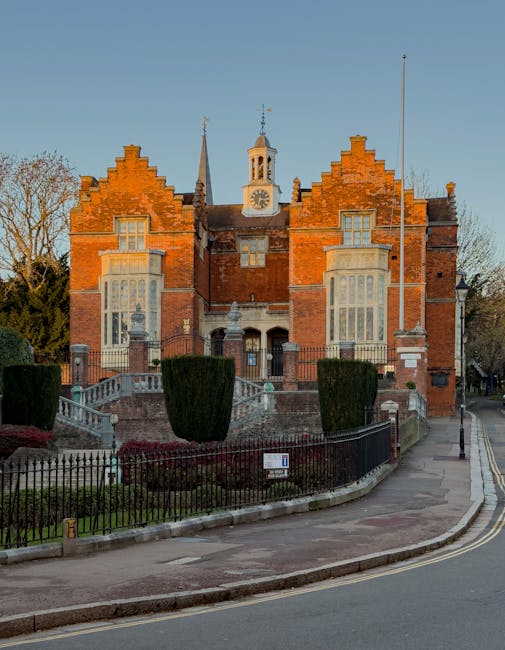 A large historic red brick building with Gothic architectural features, including pointed gables and tall windows, situated on a curved street in daylight. In the foreground, a black metal fence surro